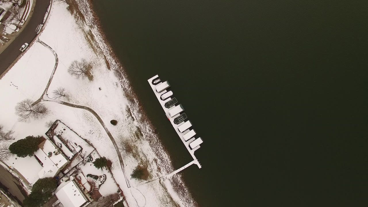 Opening overhead frame in Jefferson County, Colorado showing dark lake water, a snow-lined shore, curved paths, lakeside buildings, and a white dock with no visible people.