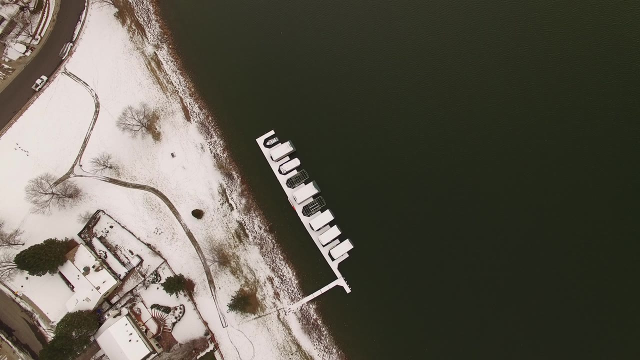 Aerial view of dark lake water beside snowy ground, homes, and a dock.