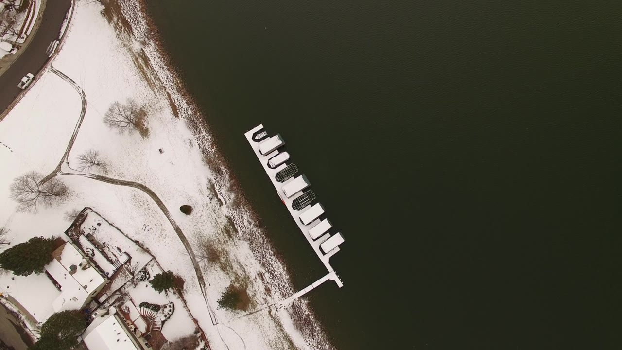 Aerial view of snowy shore and dock beside dark water.