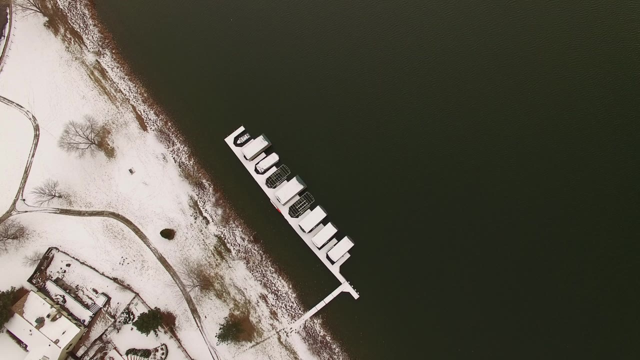 Overhead aerial frame in Jefferson, Colorado showing dark lake water, a snow-covered shore, a white dock with small boats or slips, curved paths, bare trees, and no visible people.