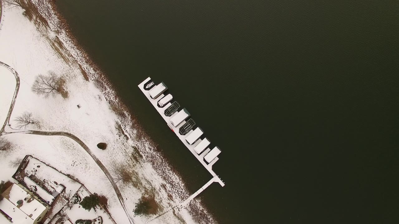 Top-down winter lake view with dock, boats, snow, and bare trees along shore.
