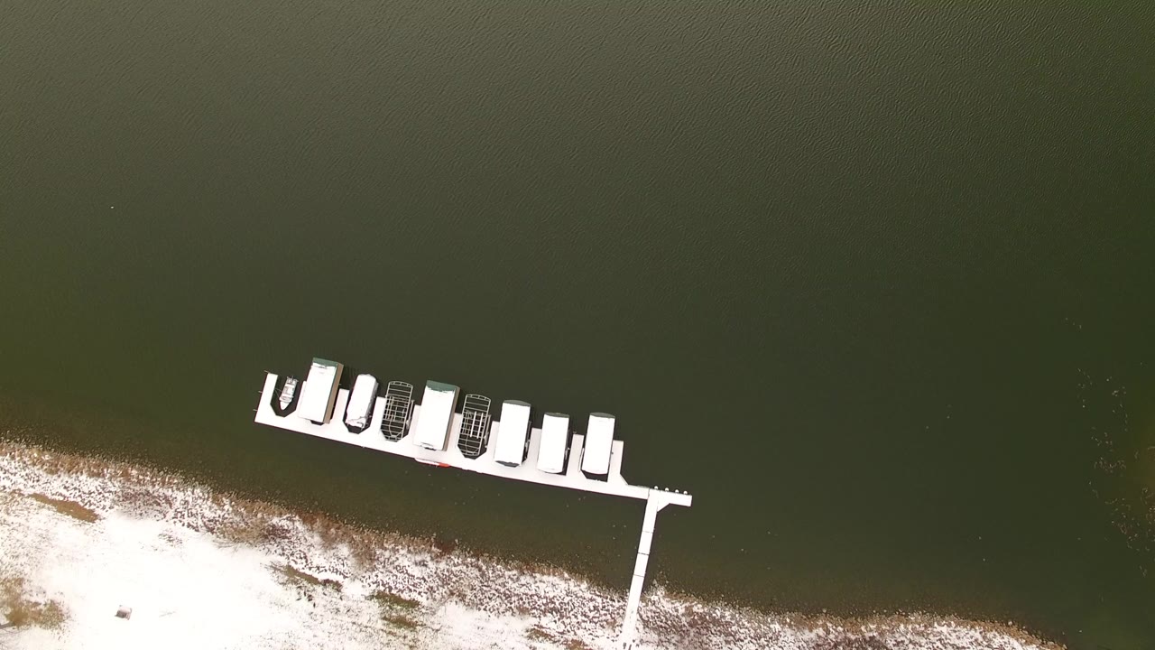 Overhead view in Jefferson, Colorado, showing a white dock with several small boats near dark green water and a snow-lined shore, with no visible people.