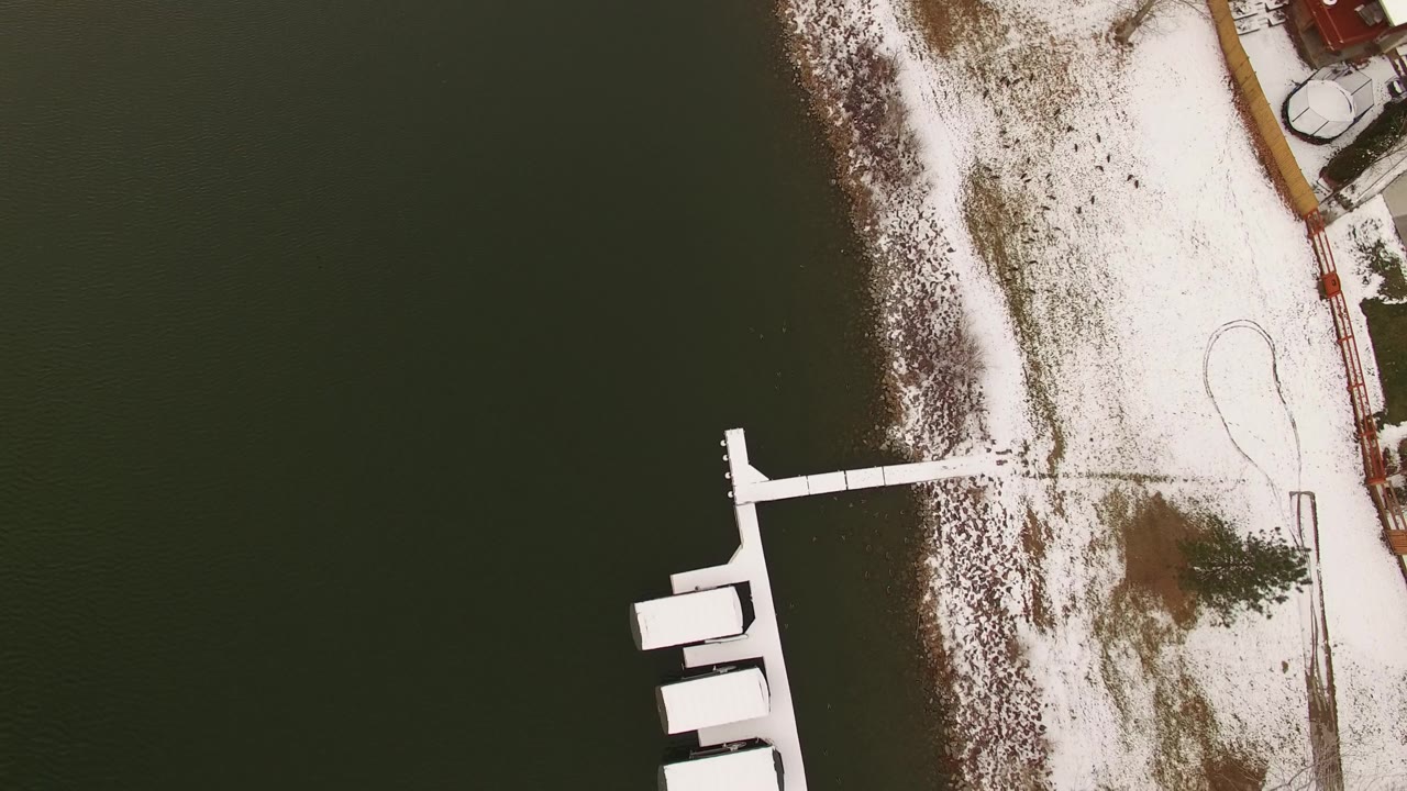 Overhead Jefferson, Colorado lake frame with a curved snow-covered bank, a white dock reaching into dark water, and a small evergreen near the shore, with no visible people.