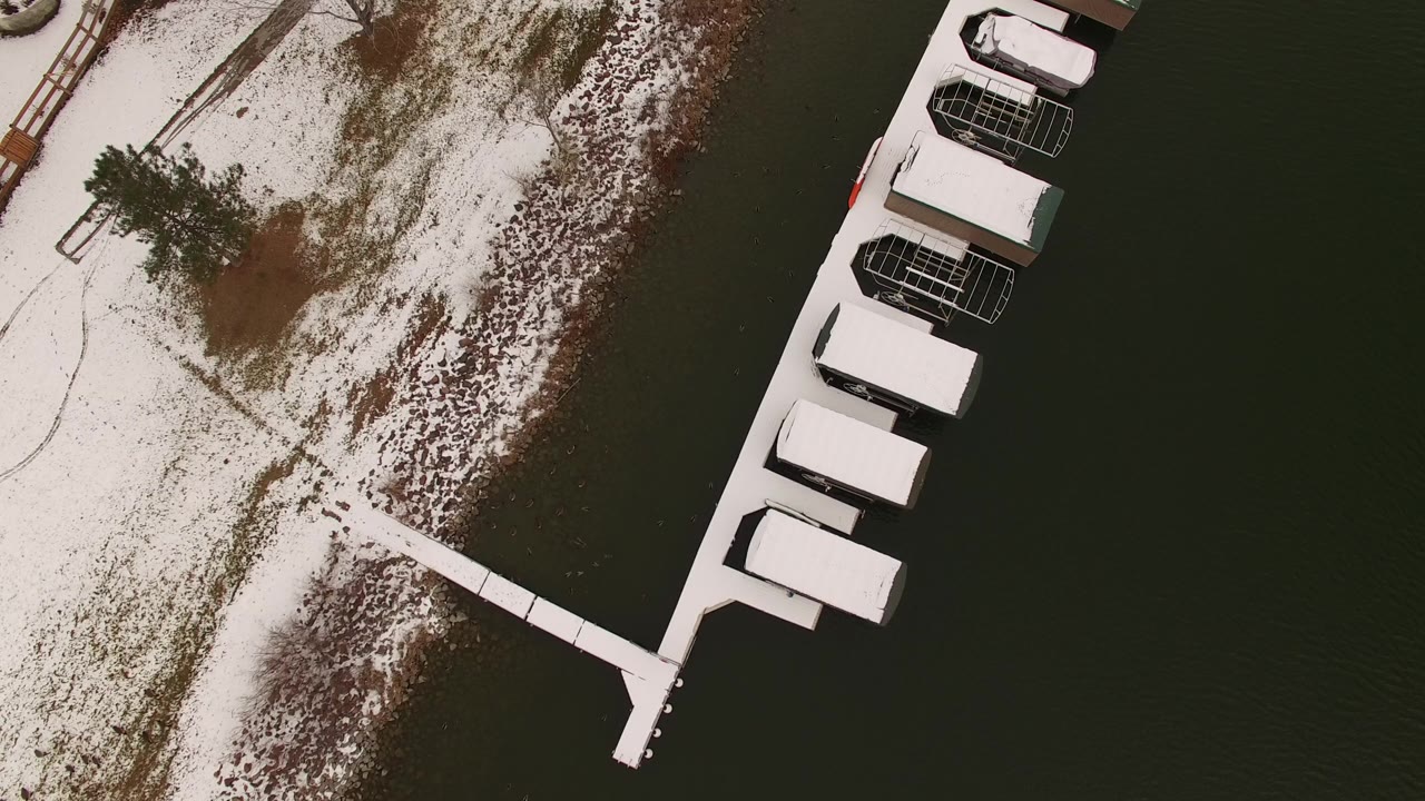 Aerial frame over Jefferson County, Colorado showing a snow-dusted shoreline, a narrow dock, dark water, and roofed lake structures along the right edge, with no visible people.