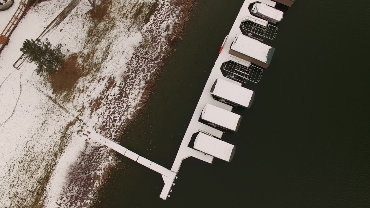 Aerial frame over Jefferson County, Colorado with patchy snow on land, a dock extending across dark water, and several snow-topped roofed structures at the right, with no visible people.