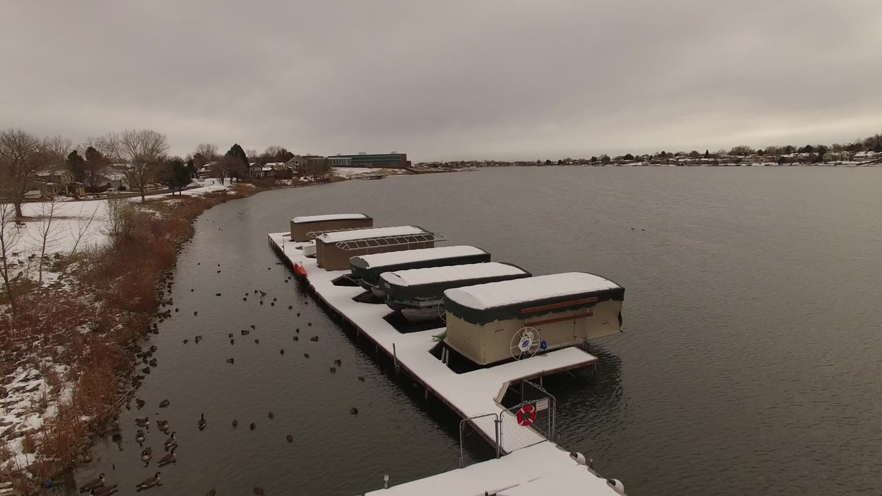 Jefferson, Colorado lake frame showing a diagonal row of snow-topped boats at the dock, open water to the right, shoreline homes in the distance, and no visible people.