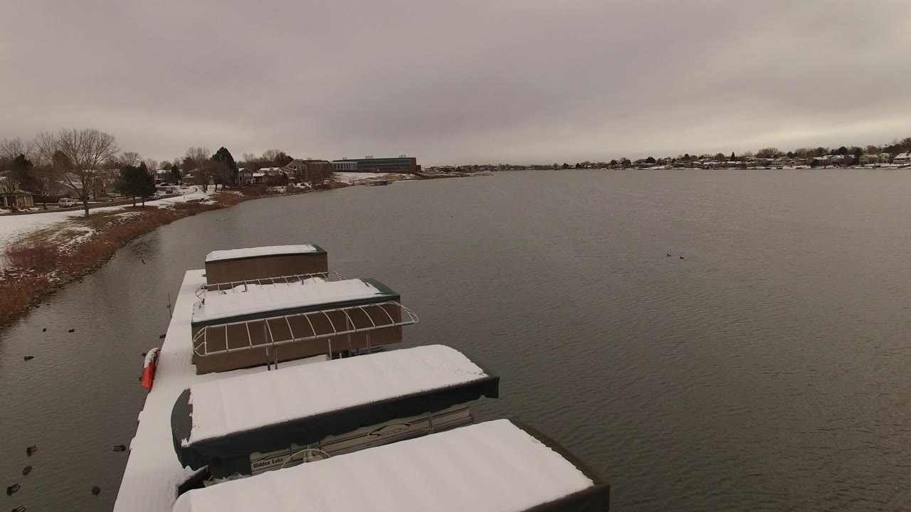 Opening view in Jefferson County, Colorado with snow-covered boat roofs at the lower frame, gray lake water beyond, bare shoreline trees, and no visible people.