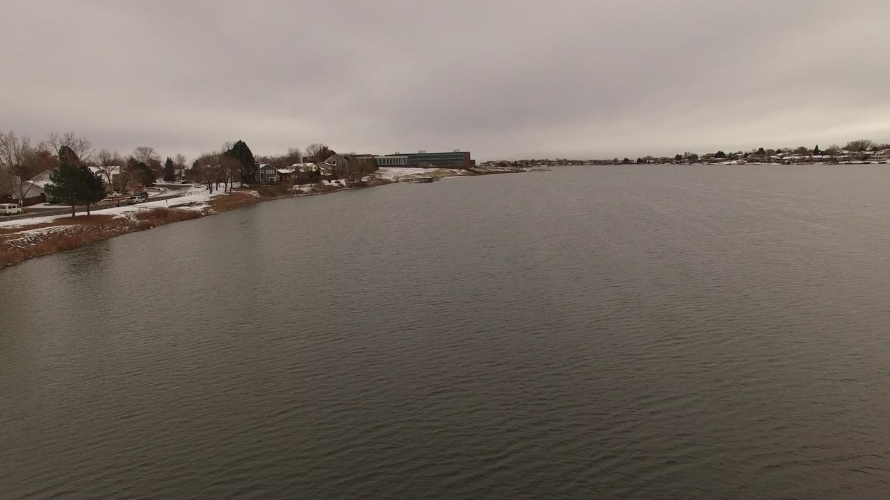 Wide water view in Jefferson County, Colorado, with ripples in the foreground, snow along the left shoreline, houses and leafless trees beyond, and no visible people.