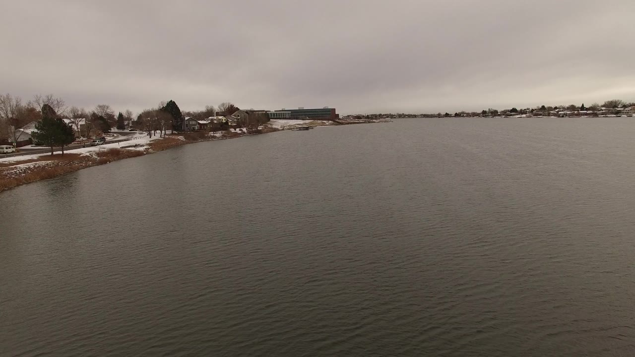 Jefferson County, Colorado waterway with a snowy bank at left, clustered homes and bare trees near the shore, open gray water across the frame, and no visible people.