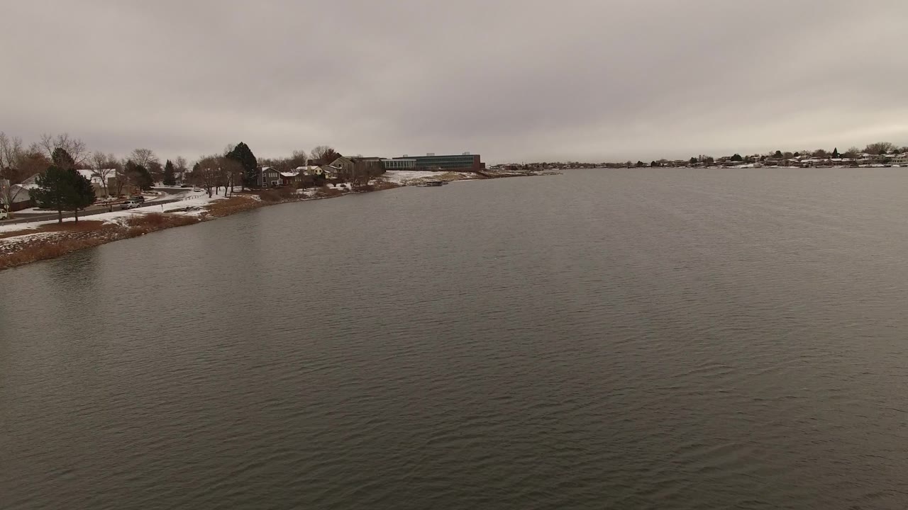 Gray water with snowy bank, trees, homes, and distant building