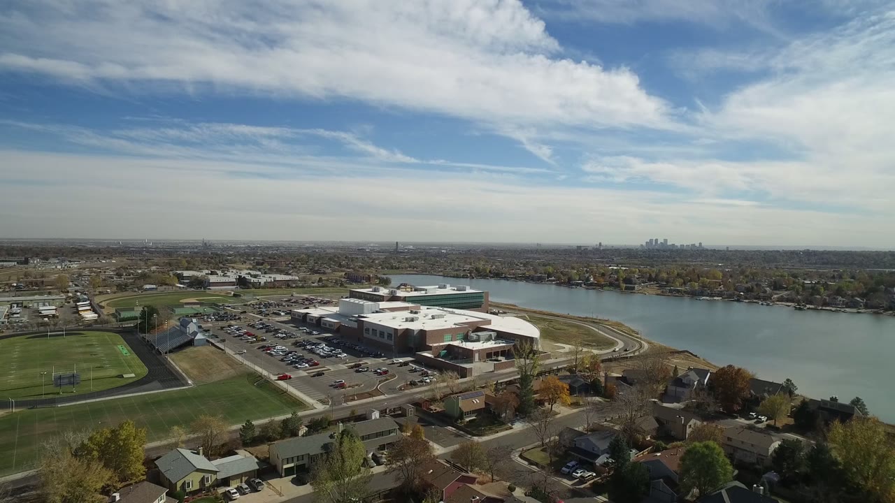 Wide aerial frame in Hidden Lake, Colorado showing a white-roofed office building by water, green athletic fields, residential streets, distant horizon, and no visible people.