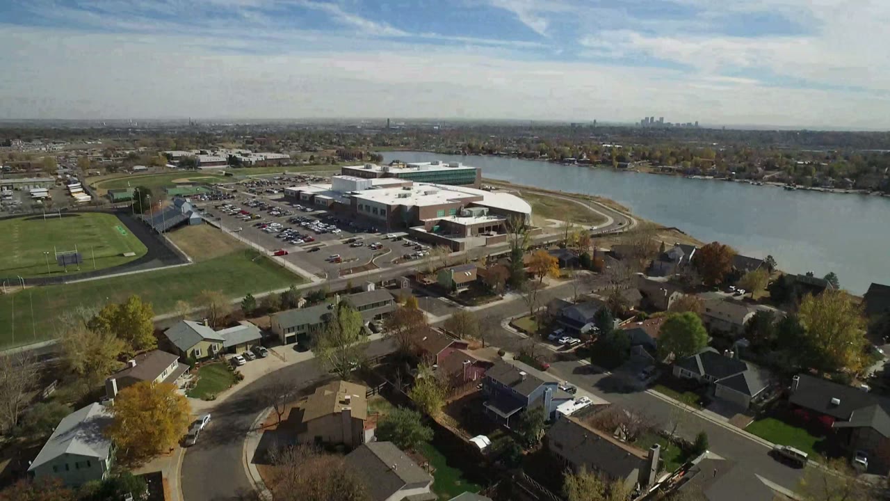 Aerial frame in Hidden Lake, Colorado looking over detached homes and autumn trees toward a parking lot, office building, athletic field, water, and no visible people.