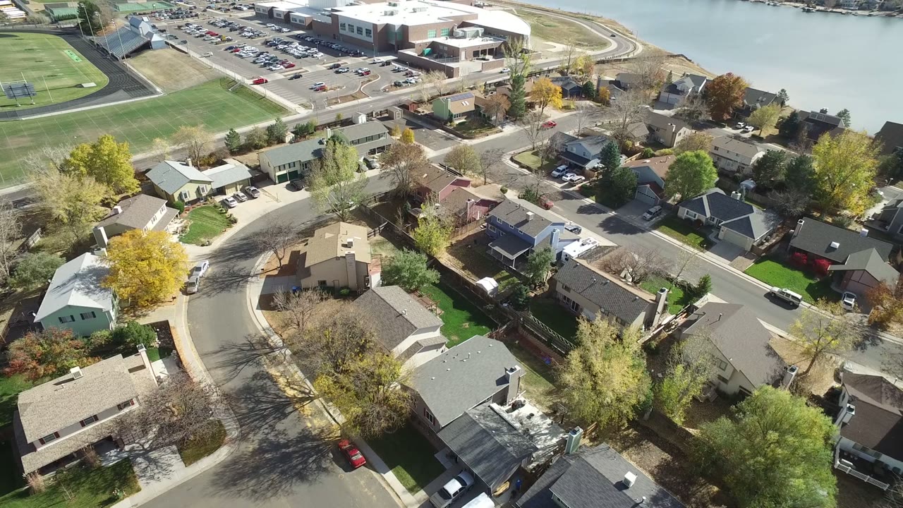 Aerial view of suburban houses, streets, trees, parking, sports fields, and water.