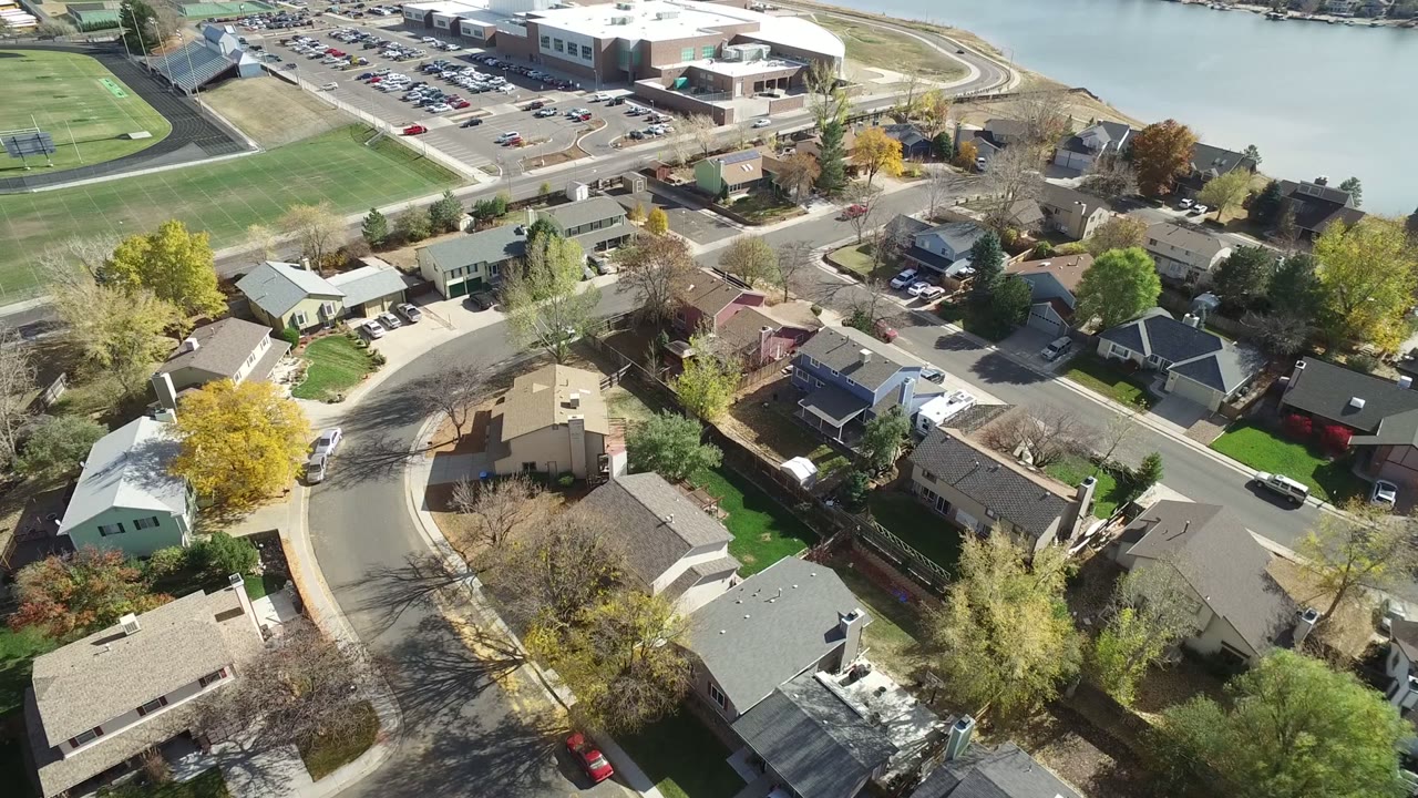 Aerial frame from Hidden Lake, Colorado, centered on suburban roofs and a curved street, with green athletic fields, parked cars, and lake water in the background, no visible people.