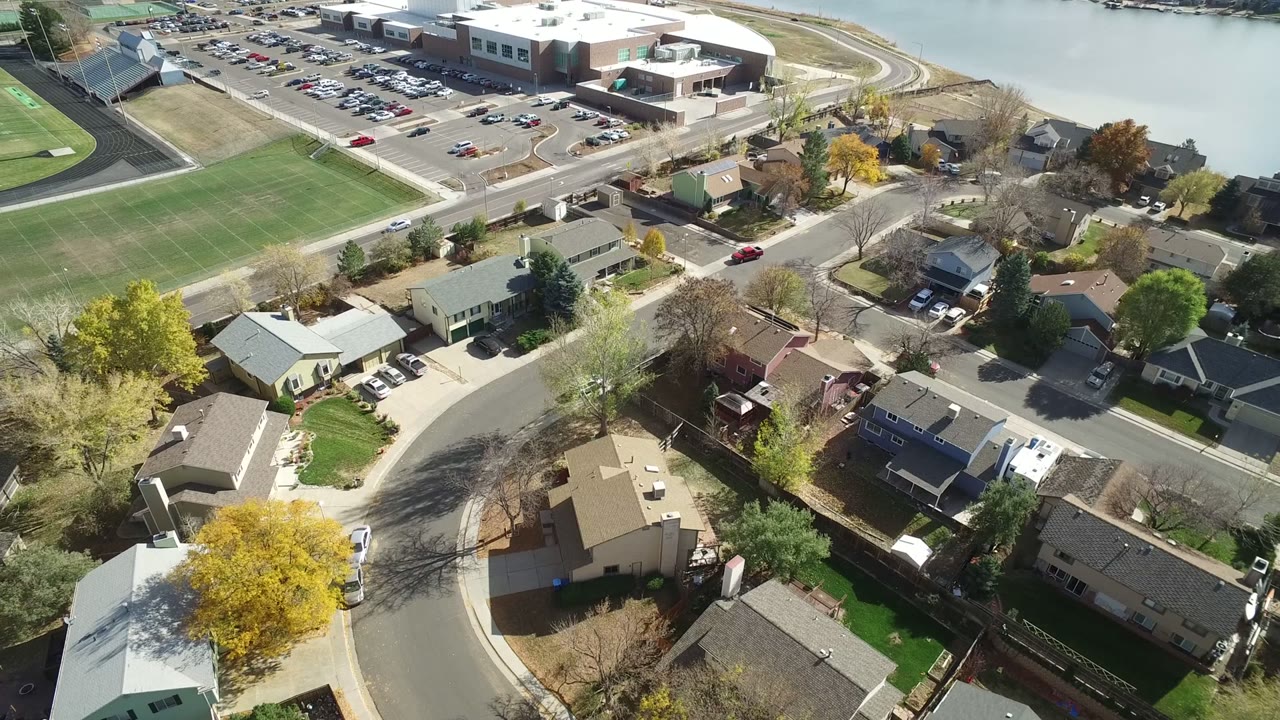 Aerial frame from Hidden Lake, Colorado, showing tan and gray rooftops around a curved street, fall-colored trees, parked cars, the green field and a large parking lot in bright sun, with no visible people.