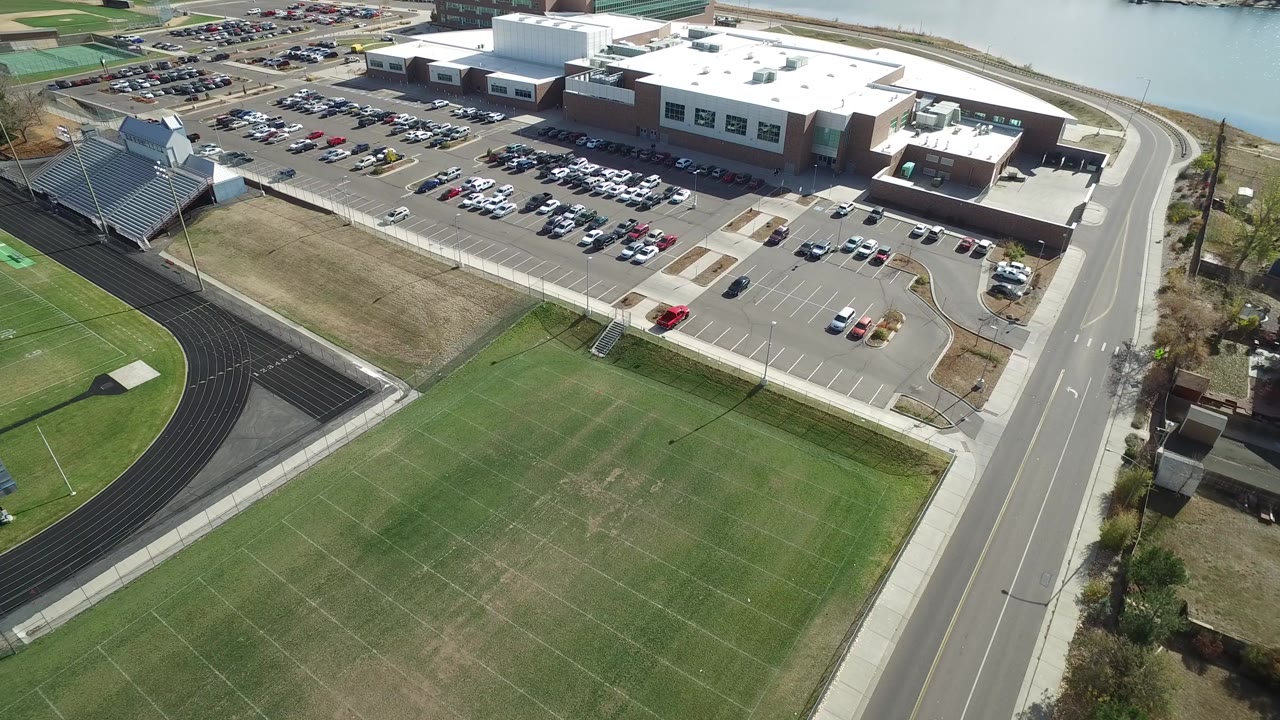 Opening aerial frame over Hidden Lake, Colorado, showing green sports fields, a black running track, bleachers, a large building, parking lots, a road, and no visible people.