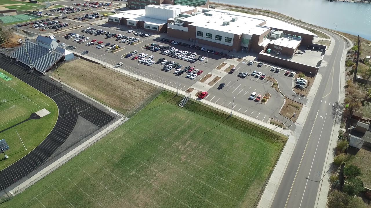 Drone-height view of a running track, sports fields, building complex, parking rows, and road.