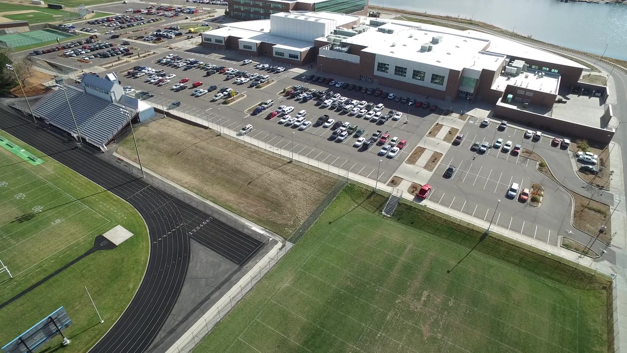 High aerial frame over Hidden Lake, Colorado showing a large white-roofed building, crowded parking lots, metal bleachers, a black track, and a green football field, with no visible identifiable people.