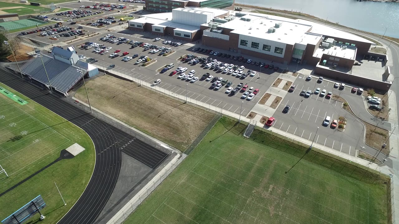 Aerial frame in Hidden Lake, Colorado with the black track curving through the foreground, bleachers at left, parked cars around a large building, and water visible beyond the roofline.
