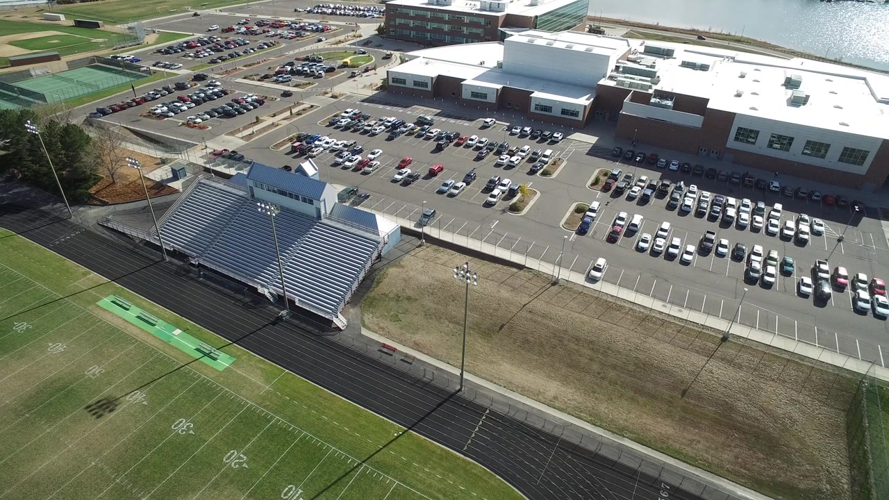 Aerial frame over Hidden Lake, Colorado showing metal bleachers and a football field in the foreground, full parking lots and a large building beyond, with no visible people.