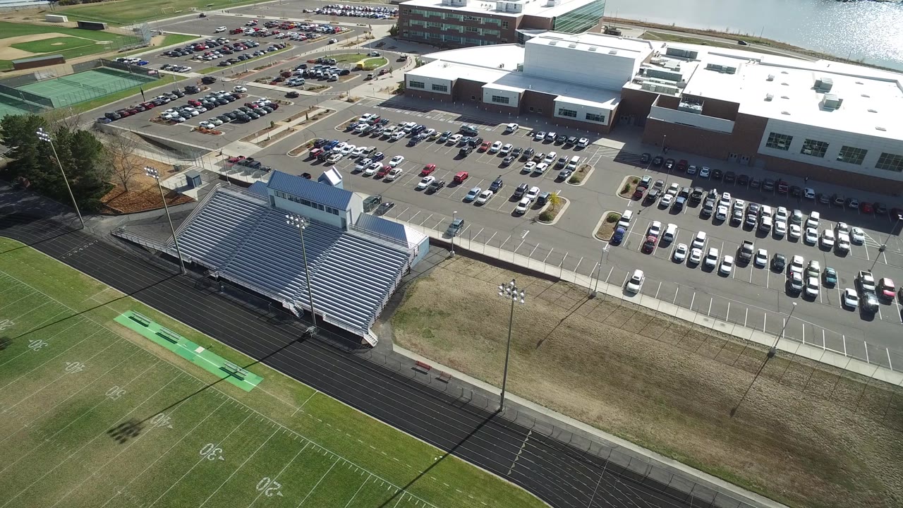 Drone-like aerial angle over a sports field, bleachers, parking lots, buildings, and water.
