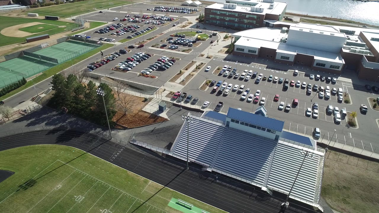 Aerial view of a football field, track, bleachers, tennis courts, parking lots, buildings, and water.