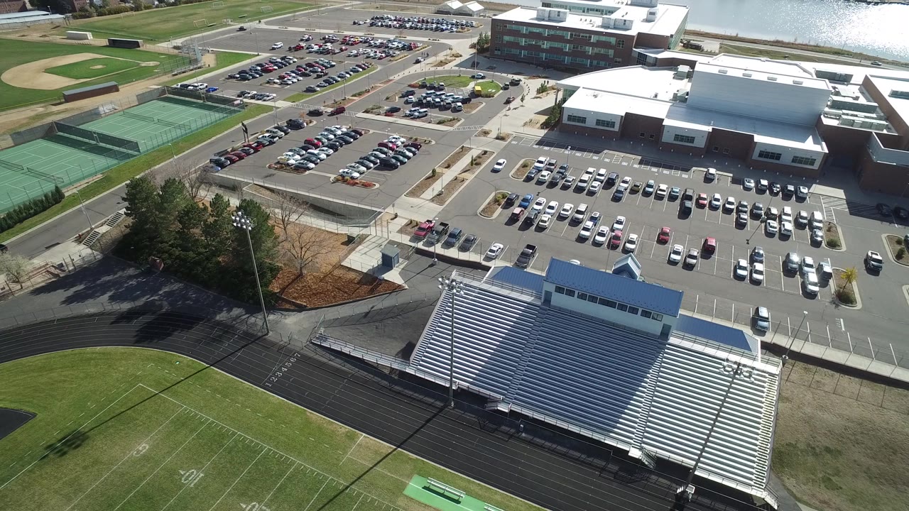 A second high-angle view in Hidden Lake, Colorado keeps the bleachers low in frame with the parking lots and tennis courts centered, water beyond the buildings, and no visible people.