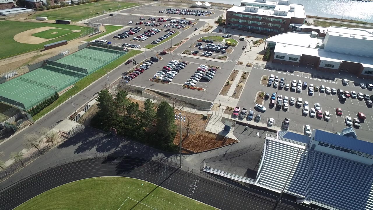Opening aerial frame in Hidden Lake, Colorado showing parking rows, green tennis courts, a track curve, bleachers, buildings, trees, roads, and tiny distant people.