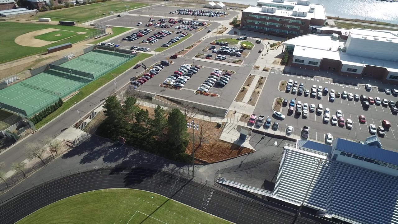 Aerial frame in Hidden Lake, Colorado with the parking rows centered, tennis courts at left, a baseball diamond near the top, bleachers at lower right, and water along the upper edge.