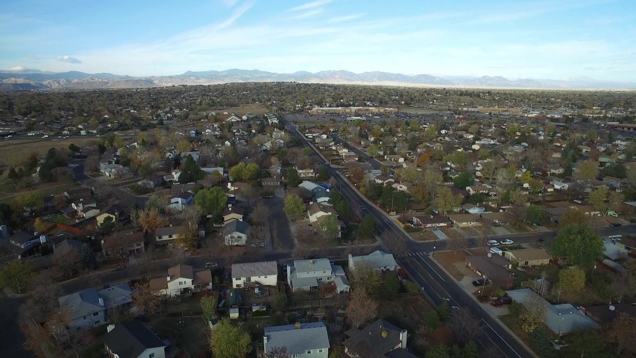 Aerial view in Hidden Lake, Colorado with a straight residential road running through houses, sparse trees, yards, parked vehicles, and mountains in the distance.