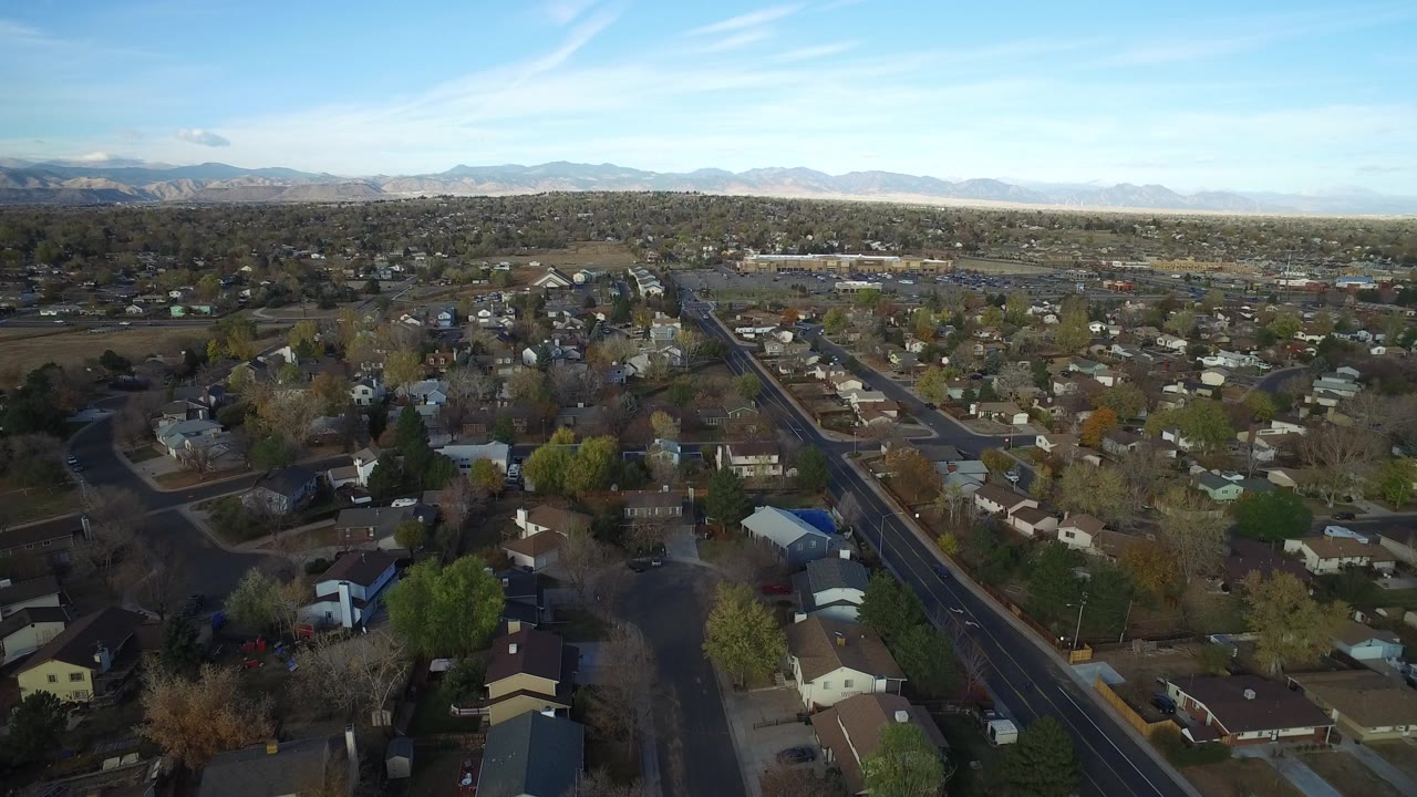 Opening aerial frame over Hidden Lake, Colorado showing residential blocks, a straight road through the neighborhood, distant mountains, blue sky, and no visible people.