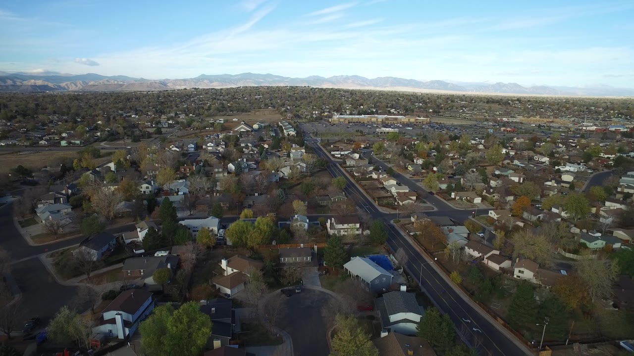 Second frame of Hidden Lake, Colorado from above, with a diagonal main road cutting past homes, trees, parked cars, and distant mountain ridges, with no visible people.