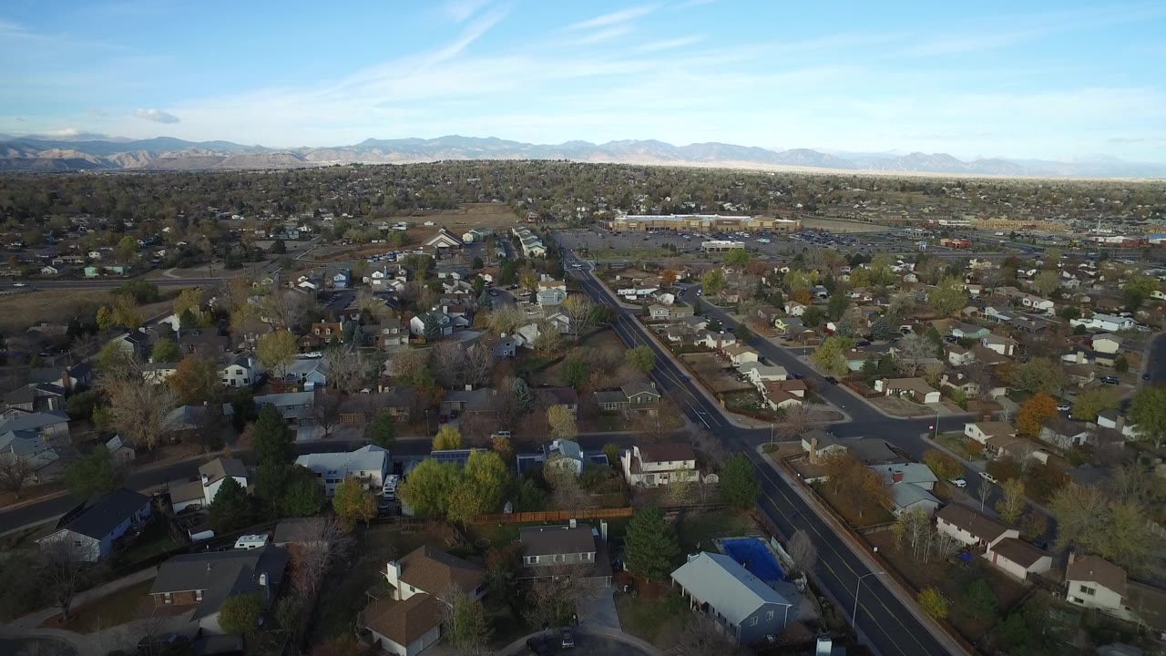 Aerial frame over Hidden Lake, Colorado, with a main road running through foreground homes, scattered trees, distant mountains, clear sky, and no visible people.