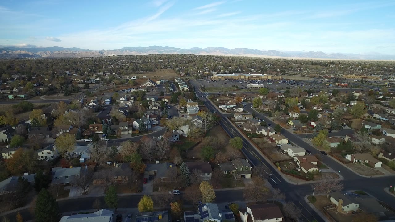 At 0:01, the Hidden Lake Colorado aerial view keeps a diagonal road prominent through the homes, with trees, open lots, distant mountains, and no visible people.