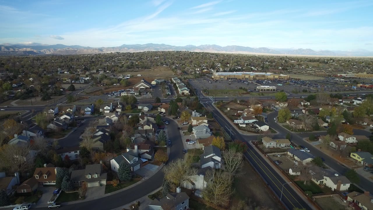 Second frame from the Hidden Lake, Colorado aerial, with rooftops and neighborhood streets below a low commercial area and pale mountains in the distance.