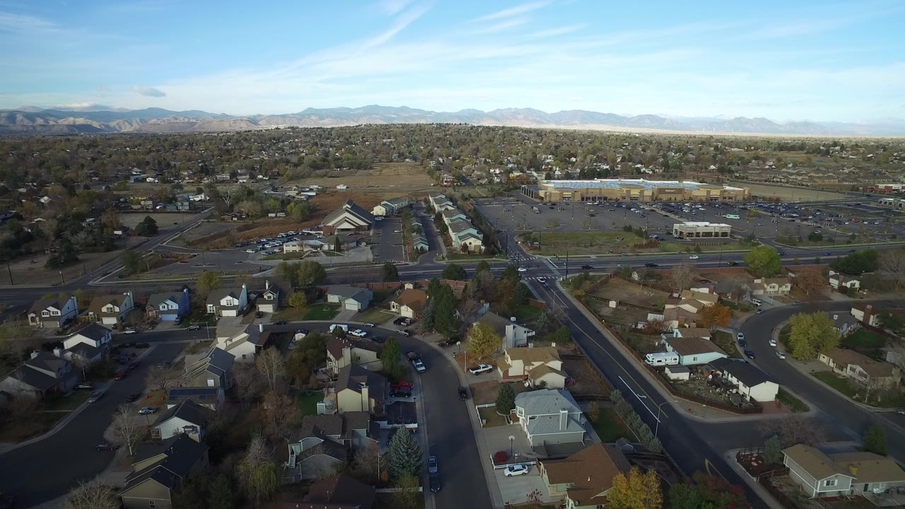 Aerial frame over Hidden Lake, Colorado showing foreground houses, a main road, commercial parking areas, distant mountains, blue sky, and no visible people.