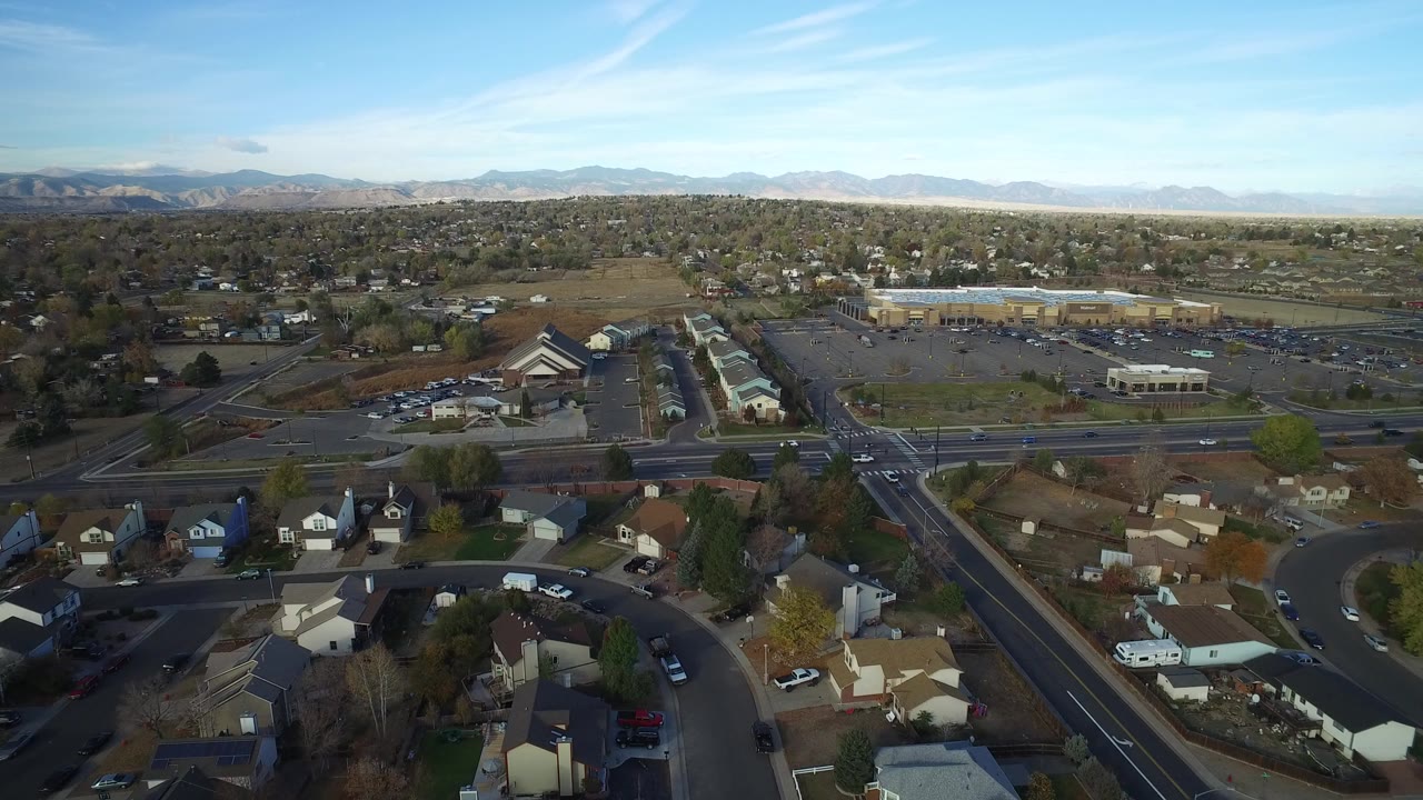 Aerial view over suburban homes, a wide road, retail parking lots, and distant mountains.