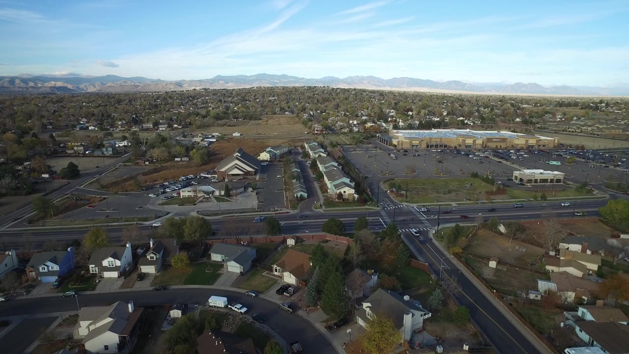Aerial frame over Hidden Lake, Colorado, with the main intersection centered below, homes along the lower edge, business parking lots in the midground, mountains on the horizon, and no visible people.