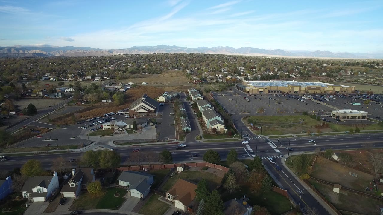 Opening aerial frame over Hidden Lake, Colorado with houses in the foreground, a multi-lane road and parking lots beyond, distant mountains under a blue sky, and no visible people.