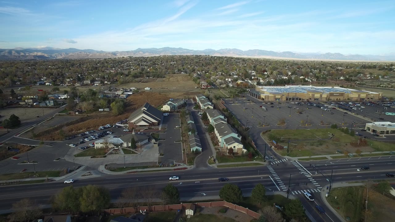 Opening aerial frame over Hidden Lake, Colorado, with a wide highway and crosswalk in front, low buildings and houses beyond, mountains on the horizon, and no visible people.