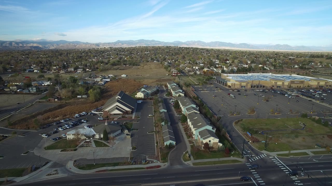 Aerial view in Hidden Lake, Colorado, with long light-roofed buildings in the foreground, a large parking lot to the right, houses across the midground, mountains on the horizon, and no visible people.