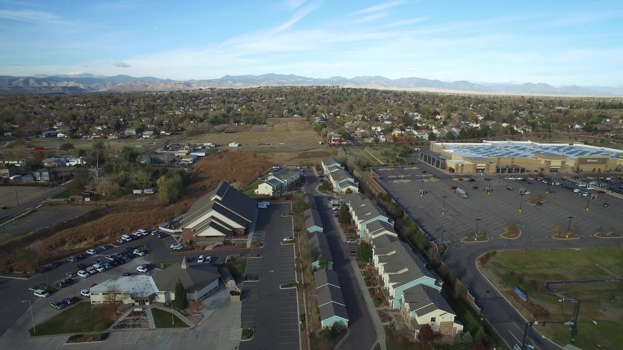 Aerial view in Hidden Lake, Colorado with a church-like roof in the lower frame, townhouses along a road, a large parking lot, homes, mountains, and no visible people.