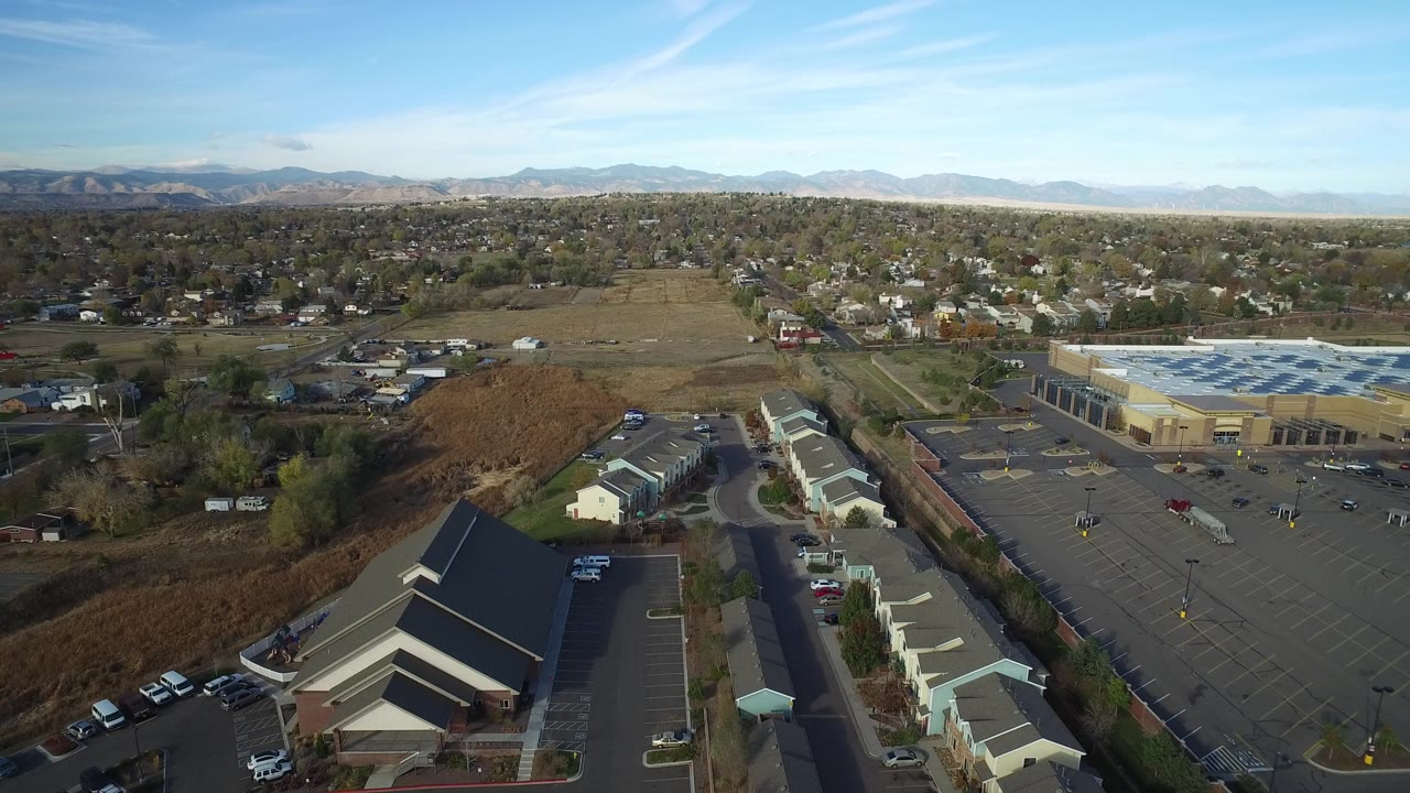Opening aerial frame over Hidden Lake, Colorado showing a parking lot, low commercial buildings, nearby homes, open fields, distant mountains, and no visible people.