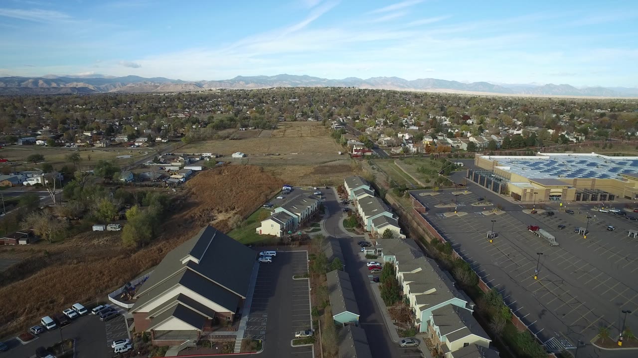 Second aerial frame of Hidden Lake, Colorado with a dark-roofed building in the foreground, a mostly empty retail lot to the right, homes beyond it, and no visible people.