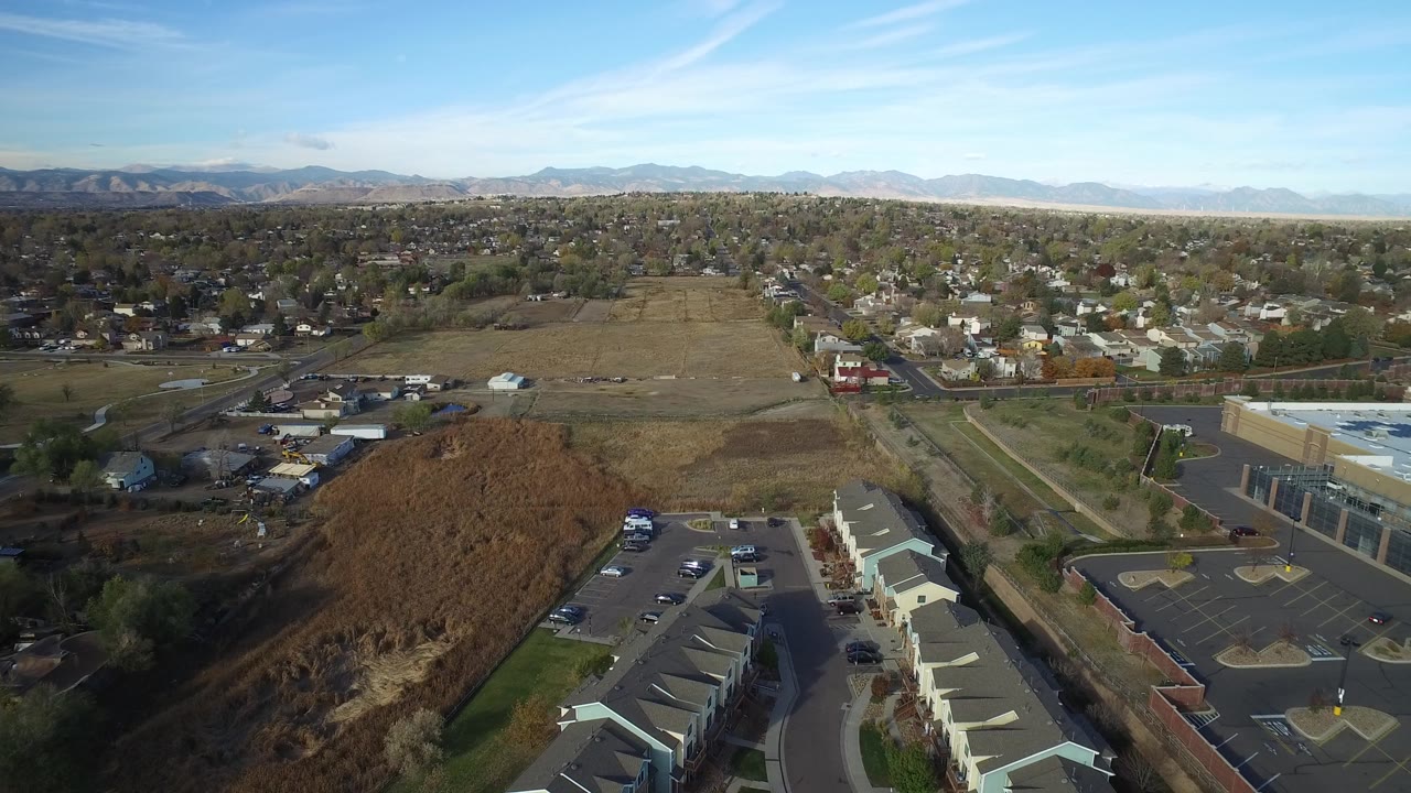 Aerial view in Hidden Lake, Colorado with an open field centered between residential blocks, townhouse roofs and parked cars below, foothills on the horizon, and no visible people.