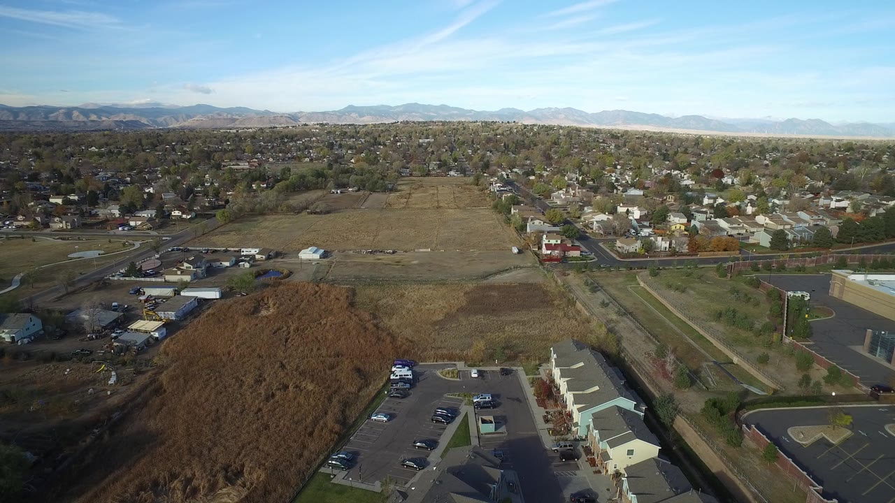 Aerial frame over Hidden Lake, Colorado showing open brown fields in the foreground, suburban homes beyond, distant mountains, blue sky, and no visible people.