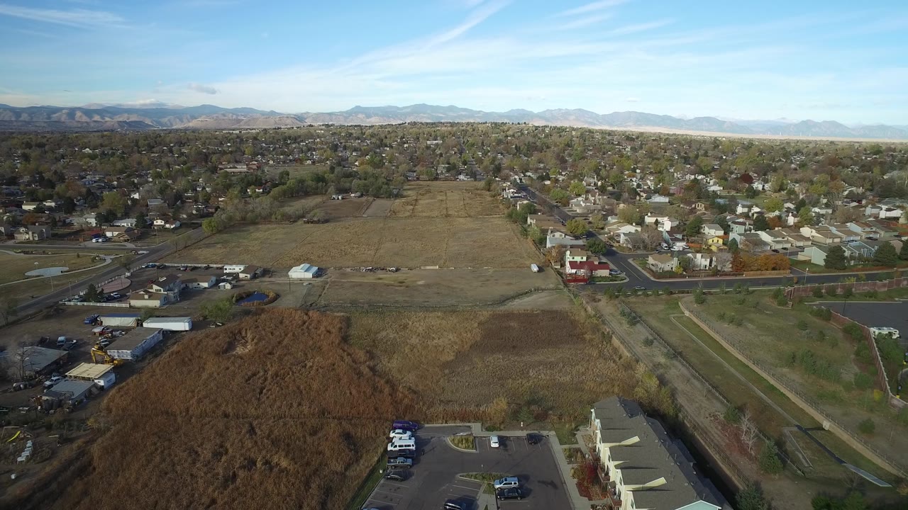 Early aerial frame over Hidden Lake, Colorado, showing brown field areas, a road corridor, compact neighborhoods, distant mountains, and no visible people.