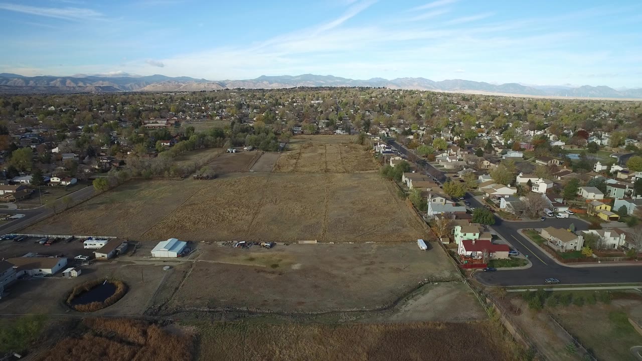 Opening aerial frame over Hidden Lake, Colorado, with a large dry undeveloped plot centered beside neighborhood streets, homes, a small pond, distant mountains, and no visible people.