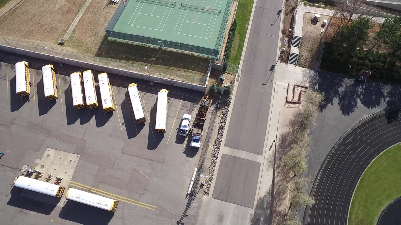 Opening overhead frame in Hidden Lake, Colorado showing rows of buses on gray tarmac, green tennis courts at the top, a road at right, and no visible people.