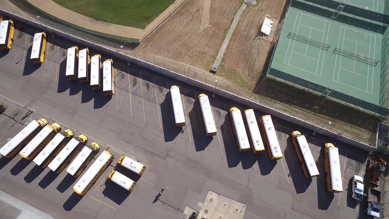 Overhead frame from Hidden Lake, Colorado shows rows of yellow-and-white buses on gray pavement beside green tennis courts, with no visible people in this opening moment.